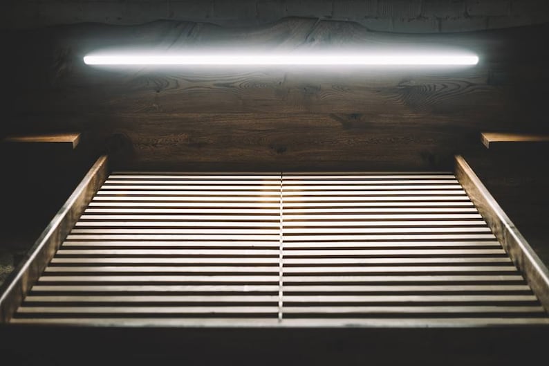 May include: A dark, moody image of a wooden bed frame with a slatted base. Above the bed, a wooden headboard and a bright, linear light fixture are visible. The scene is dimly lit, creating a sense of calm.