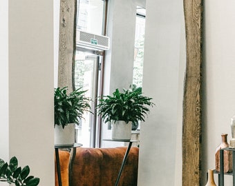 Op de afbeelding: Grote rechthoekige spiegel met een natuurlijke houten lijst. De spiegel weerspiegelt een kamer met planten, een bank en een raam. De lijst heeft een ruwe, onafgewerkte uitstraling, wat een vleugje organische stijl toevoegt. Een meubelstuk.