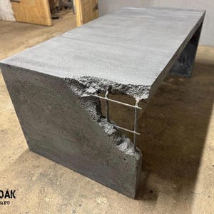 Op de afbeelding: Een grijze betonnen bank met een rechthoekige bovenkant en een gedeeltelijk blootgestelde binnenkant met metalen wapening. De bank heeft een moderne, industriële uitstraling. Het OAKY OAK furniture logo is zichtbaar.