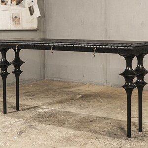 May include: A black console table with a distinctive design. The table has a rectangular top with two drawers and ornate legs. The legs have a unique shape, with multiple bulbous sections. The table is made of wood and has a matte finish. The table's dimensions are in centimeters.