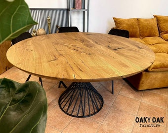 May include: A round wooden dining table with a natural oak finish. The table has a black metal base with a unique, geometric design. The table is surrounded by black chairs and a mustard-colored sofa. The table is from Oaky Oak Furniture.