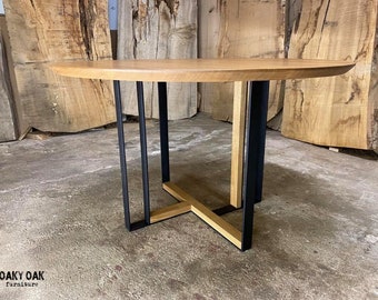 May include: A round wooden table with a light brown top and black metal legs. The legs have a unique design, with a cross-shaped base. The table is a modern piece of furniture.