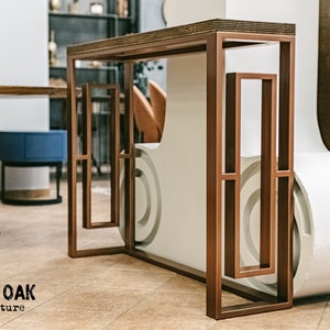 Könnte beinhalten: Ein moderner Konsolentisch aus braunem Metall mit geometrischem Design. Der Tisch hat eine rechteckige Platte und einen Rahmen mit vertikalen und horizontalen Linien. Der Tisch steht in einem Ausstellungsraum mit anderen Möbeln.