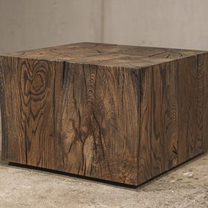 May include: A solid, dark brown wooden cube-shaped coffee table. The wood grain is visible, with natural cracks and variations in the surface. The table has a modern, minimalist design and sits directly on the floor.
