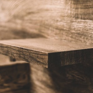 May include: Close-up of a floating wooden shelf with a dark brown finish. The shelf is rectangular and appears to be made of solid wood, with visible grain and texture. The background is a wooden wall.