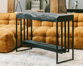 Puede incluir: Una consola moderna con una tapa de madera oscura y texturizada y una estructura de metal negro. La mesa tiene un estante inferior y está sostenida por barras metálicas verticales. La madera parece toscamente labrada, dándole una estética rústica.