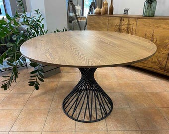 May include: Round wooden dining table with a light brown finish. The table has a black, spiral-shaped metal base. The table is set on a tiled floor. The overall style is modern and minimalist.