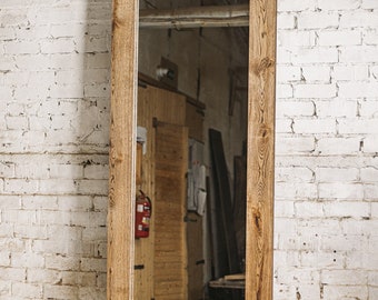 Op de afbeelding: Volledige spiegel met een rechthoekig reflecterend oppervlak omlijst door twee verticale houten planken. Het hout heeft een natuurlijk nerfpatroon en een warme, lichtbruine kleur. De spiegel staat tegen een witte bakstenen muur.