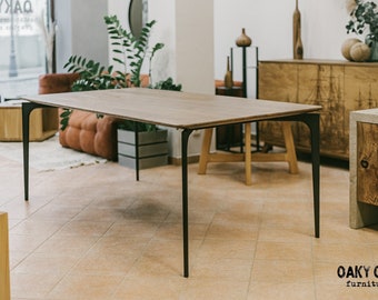 May include: A rectangular wooden table with a light brown surface and black metal legs. The table is in a showroom setting with other furniture and plants. The brand name "Oaky Oak Furniture" is visible.