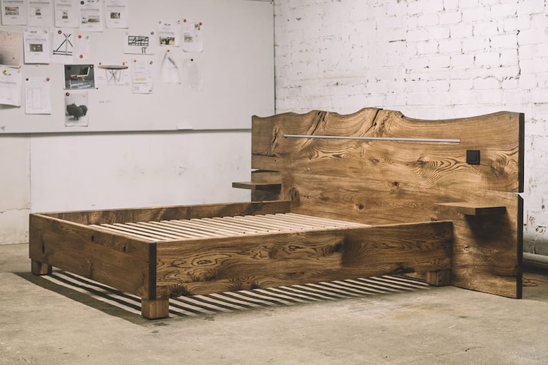 May include: A handcrafted wooden bed frame with a natural, rustic aesthetic. The headboard features a unique, organic edge and a horizontal metal bar. The bed includes integrated side tables and a slatted base for the mattress. The wood has a warm, natural tone.