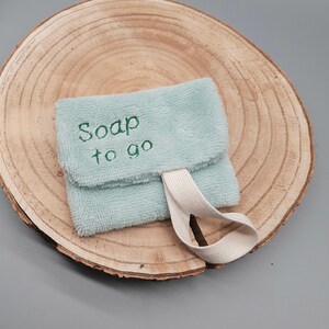 Könnte beinhalten: Ein hellblauer Seifensäckchen aus Stoff mit einem weißen Gummiband. Das Säckchen hat den Text "Soap to go" auf der Vorderseite gestickt.
