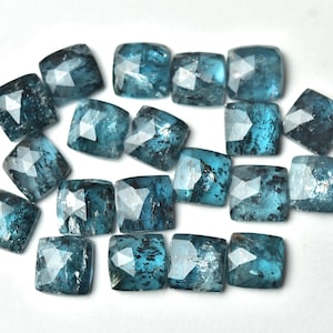 May include: A collection of square-cut, teal-colored gemstones with faceted surfaces. The gems have a slightly mottled appearance and are arranged on a white background. These stones could be used for jewelry making or other crafts.