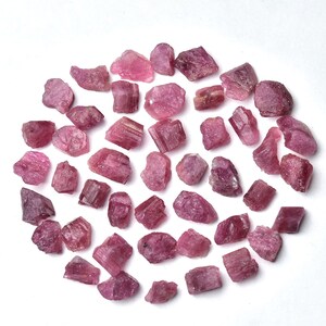 May include: A collection of raw, rough-cut pink tourmaline gemstones. The stones vary in size and shape, with a range of pink hues. The gemstones are scattered on a white surface, creating a visually appealing arrangement.