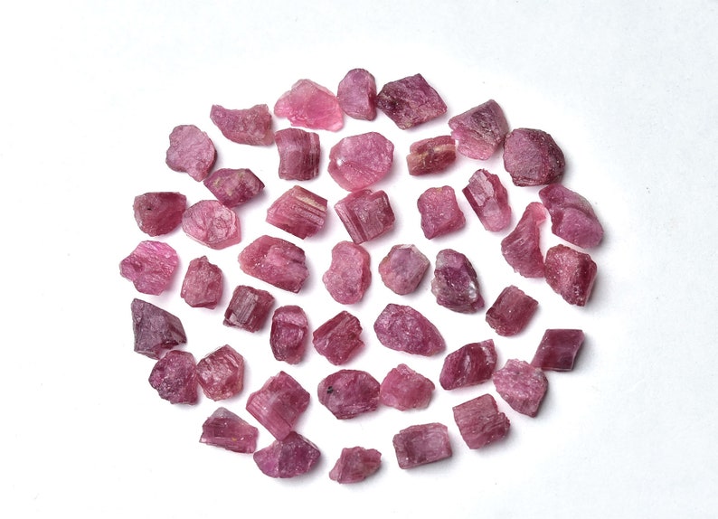 May include: A collection of raw, rough-cut pink tourmaline gemstones. The stones vary in size and shape, with a range of pink hues. The gemstones are scattered on a white surface, creating a visually appealing arrangement.