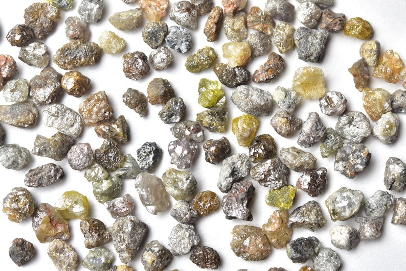 May include: A collection of small, rough, gray and yellow diamonds. The diamonds are scattered on a white surface.