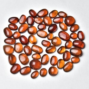 May include: A collection of approximately 40 smooth, oval-shaped, reddish-brown gemstones. The stones are arranged in a loose circle on a white background.