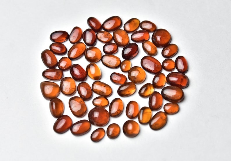 May include: A collection of approximately 40 smooth, oval-shaped, reddish-brown gemstones. The stones are arranged in a loose circle on a white background.