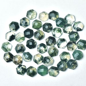 May include: A collection of faceted, hexagonal gemstones with a moss agate appearance. The stones are primarily green with white and darker green inclusions, arranged on a white surface. The gemstones have a polished, reflective surface.