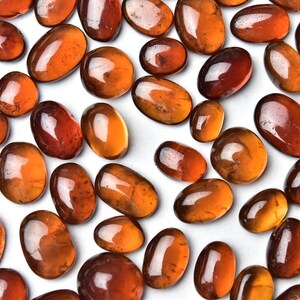 May include: A collection of smooth, polished, oval-shaped, orange-brown gemstones. The stones are arranged on a white surface.