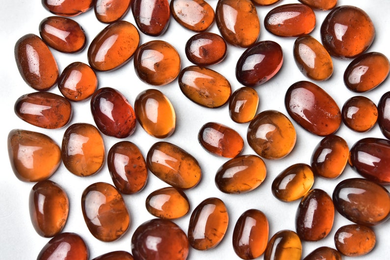 May include: A collection of smooth, polished, oval-shaped, orange-brown gemstones. The stones are arranged on a white surface.
