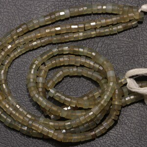 May include: A strand of light green gemstone beads with a square shape. The beads are strung on a white cord with tassels at the ends.