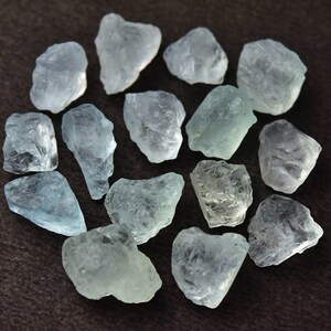 Puede incluir: Un conjunto de piedras preciosas de aguamarina azul claro en bruto. Los cristales en bruto y sin cortar var&iacute;an en tama&ntilde;o y forma, mostrando una calidad transl&uacute;cida. Las piedras est&aacute;n dispuestas sobre una superficie marr&oacute;n oscuro, mostrando su belleza natural.