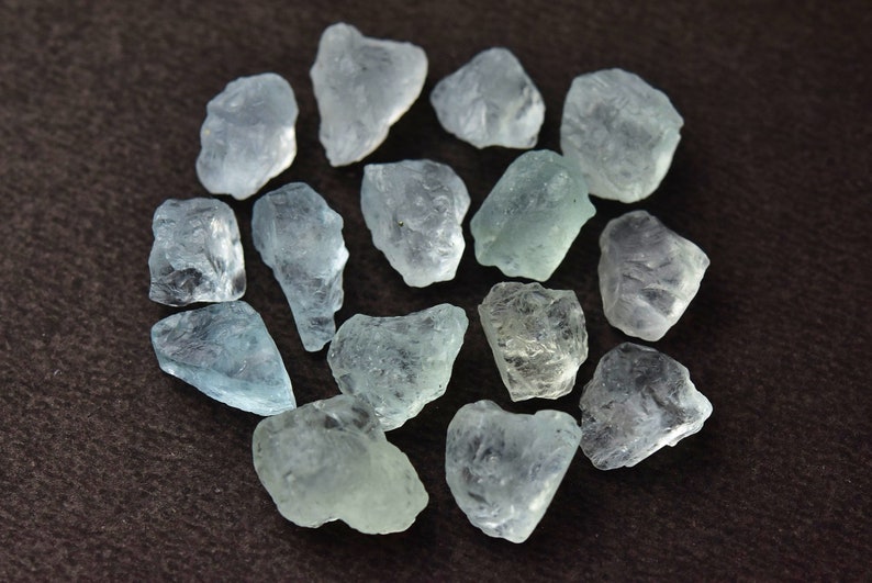 Puede incluir: Un conjunto de piedras preciosas de aguamarina azul claro en bruto. Los cristales en bruto y sin cortar var&iacute;an en tama&ntilde;o y forma, mostrando una calidad transl&uacute;cida. Las piedras est&aacute;n dispuestas sobre una superficie marr&oacute;n oscuro, mostrando su belleza natural.