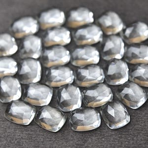 May include: A collection of faceted, silver-colored gemstones arranged in a circular pattern. Each gem has a multifaceted surface, reflecting light. The gems are set against a dark gray background.