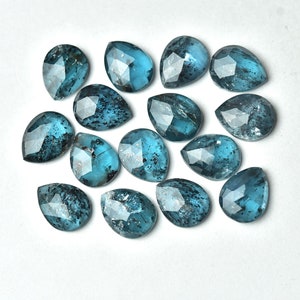 May include: A collection of teardrop-shaped gemstones with a faceted cut. The gems are a deep teal color with dark inclusions, arranged on a white surface. The gems are likely for jewelry making.