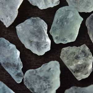 Puede incluir: Primer plano de varias piedras preciosas de aguamarina en bruto. Las piedras, de color azul verdoso p&aacute;lido, presentan una textura natural y rugosa. Los cristales sin cortar est&aacute;n dispersos sobre un fondo marr&oacute;n oscuro.