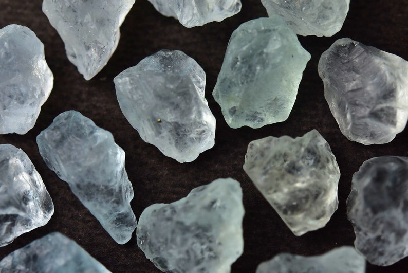 Puede incluir: Primer plano de varias piedras preciosas de aguamarina en bruto. Las piedras, de color azul verdoso p&aacute;lido, presentan una textura natural y rugosa. Los cristales sin cortar est&aacute;n dispersos sobre un fondo marr&oacute;n oscuro.