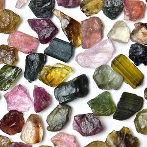 May include: A collection of rough, natural gemstones in various colors, including pink, green, brown, and black. The gemstones are scattered on a white background.