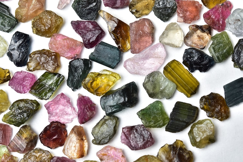 May include: A collection of rough, natural gemstones in various colors, including pink, green, brown, and black. The gemstones are scattered on a white background.