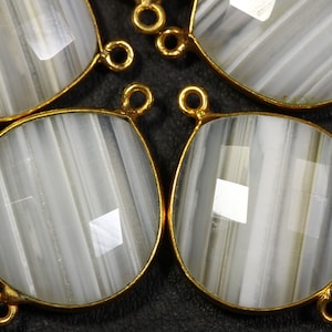 May include: Close-up of several gemstone pendants. Each pendant features a faceted, teardrop-shaped stone with gray and white banding, set in a gold-colored bezel. The pendants have small loops for attaching to jewelry.