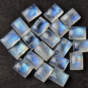 May include: A collection of 18 rectangular moonstone cabochons, each with a unique iridescent blue and white pattern. The stones are arranged in a loose cluster on a dark gray surface.