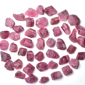 May include: A collection of raw, pink tourmaline crystals, each with a unique shape and texture. The crystals vary in size and shade, from light to deep pink, and are scattered on a white surface.