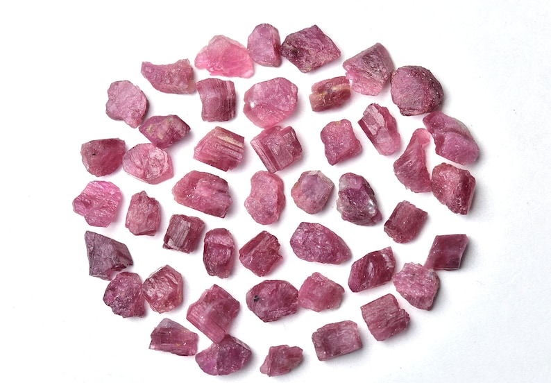 May include: A collection of raw, pink tourmaline crystals, each with a unique shape and texture. The crystals vary in size and shade, from light to deep pink, and are scattered on a white surface.