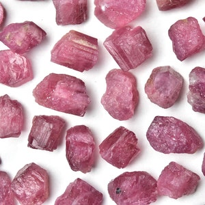 May include: A collection of raw, uncut pink tourmaline gemstones scattered on a white surface. The gemstones vary in size and shape, displaying a range of pink hues, from light to deep rose. The rough texture of the stones is clearly visible.