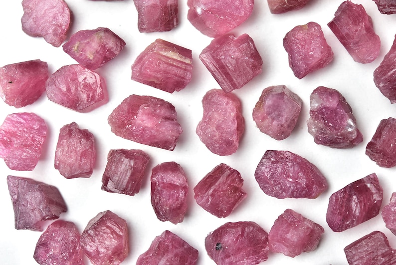 May include: A collection of raw, uncut pink tourmaline gemstones scattered on a white surface. The gemstones vary in size and shape, displaying a range of pink hues, from light to deep rose. The rough texture of the stones is clearly visible.