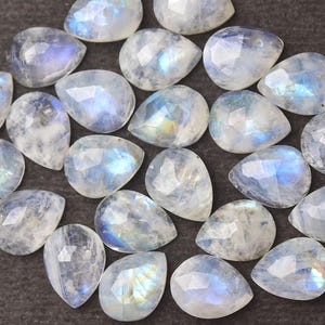 May include: A collection of faceted, pear-shaped moonstone gemstones. The stones display a milky white base with iridescent blue and yellow flashes. The gemstones are arranged on a dark gray surface, showcasing their polished surfaces and unique patterns.