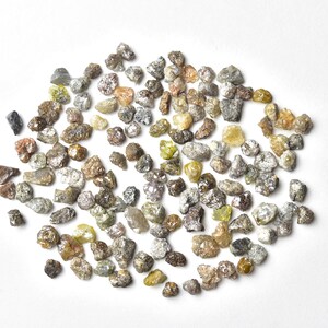 May include: A collection of rough, uncut diamonds in various shades of gray, brown, and yellow. The diamonds are scattered on a white surface.