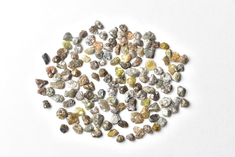 May include: A collection of rough, uncut diamonds in various shades of gray, brown, and yellow. The diamonds are scattered on a white surface.