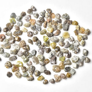 May include: A collection of small, rough, unpolished diamonds in various shades of gray, brown, and yellow. The diamonds are scattered on a white surface.
