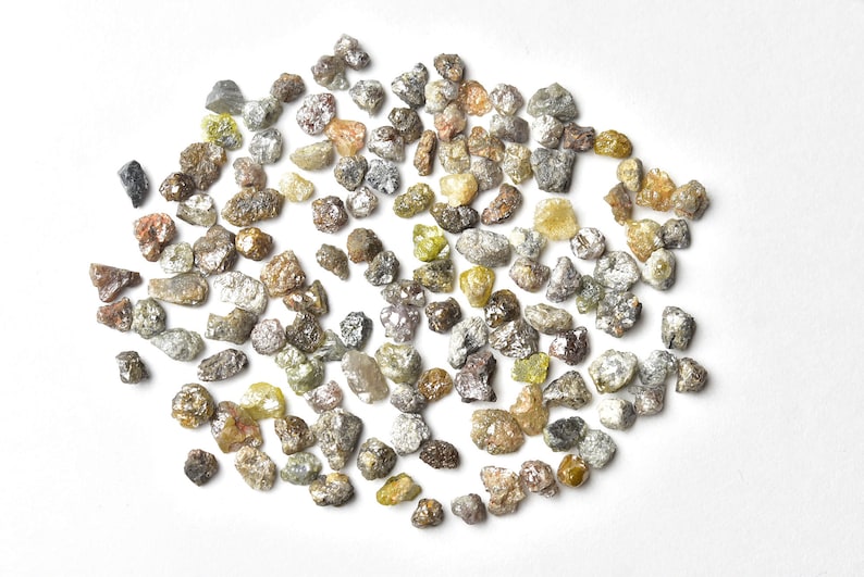 May include: A collection of small, rough, unpolished diamonds in various shades of gray, brown, and yellow. The diamonds are scattered on a white surface.