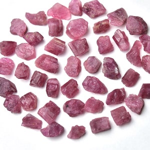 May include: A collection of raw, rough-cut pink tourmaline gemstones. The stones vary in size and shape, displaying a range of pink hues from light to deep rose. The gemstones are scattered on a white surface.