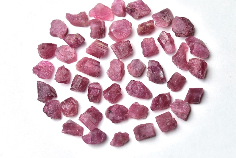 May include: A collection of raw, rough-cut pink tourmaline gemstones. The stones vary in size and shape, displaying a range of pink hues from light to deep rose. The gemstones are scattered on a white surface.