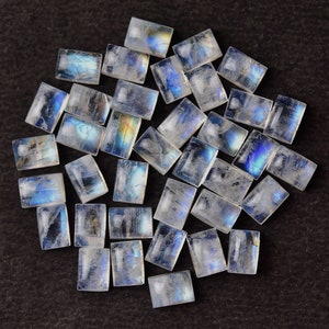 May include: A collection of iridescent rainbow moonstone cabochons in a rectangular shape. The stones are translucent and have a milky white color with a blue and green iridescent sheen.