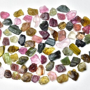 May include: A collection of rough, natural tourmaline gemstones in various colors, including green, pink, black, and yellow. The gemstones are arranged in a loose circle on a white background.