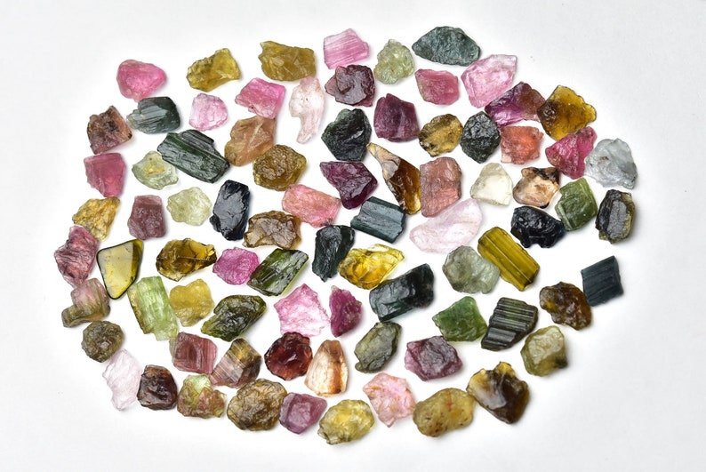 May include: A collection of rough, natural tourmaline gemstones in various colors, including green, pink, black, and yellow. The gemstones are arranged in a loose circle on a white background.