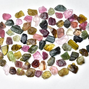 May include: A collection of rough, natural tourmaline gemstones in various colors, including green, pink, black, and yellow. The stones are arranged in a circular pattern on a white background.